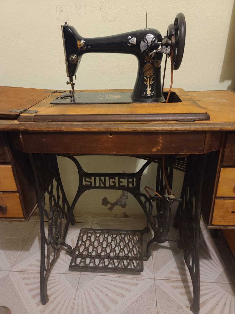 SINGERDetailed Description / Context: The Singer treadle sewing machine was a widely used household and industrial device prior to the widespread adoption of electric sewing machines. Operated by a foot-powered treadle, it allowed users to sew without electricity, making it accessible in a wide range of settings, including rural homes and small workshops. Such machines played a significant role in domestic labor, garment production, and informal economies, often used by women for household clothing production or income-generating work such as tailoring and piecework. The cabinet design, including storage drawers, reflects its integration into household furniture and daily life. The durability and widespread distribution of Singer machines made them a global standard in sewing technology, contributing to the growth of textile production and shaping labor practices in both domestic and industrial contexts.