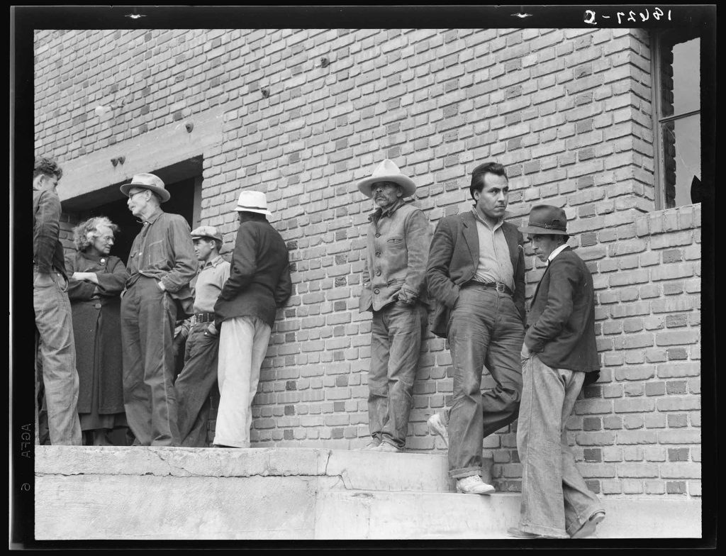Detailed Description / Context:This photograph appears consistent with images produced by federal programs such as the Farm Security Administration (FSA), which documented labor and living conditions during the 1930s and 1940s. The clothing, setting, and composition suggest a group of farmworkers, possibly migrant or seasonal laborers, gathered at a worksite, hiring location, or administrative building. The image visually complements documentary records of agricultural labor conditions in California and the broader United States, including issues of employment instability, migration, and social organization among workers. The mix of attire—ranging from work clothes to jackets and hats—may indicate different roles or statuses within the labor group. Such photographs were often used to document economic conditions, labor practices, and community life, providing visual evidence to accompany government reports and social research on agricultural workers, including those involved in programs like the Bracero Program or earlier New Deal labor initiatives.