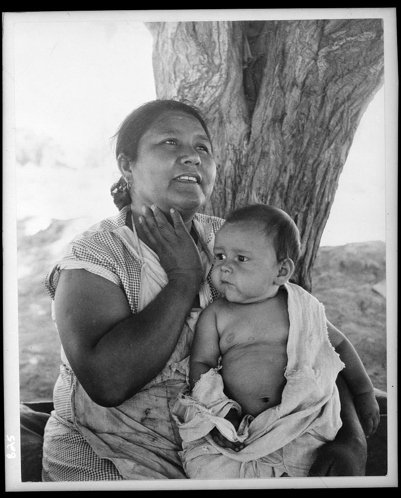 Detailed Description / Context:This photograph is consistent with documentary images produced by programs such as the Farm Security Administration (FSA), which sought to document the lives of rural and migrant populations in the United States during the 1930s and 1940s. The composition emphasizes maternal care and the presence of young children within laboring communities, suggesting the integration of family life into broader economic hardship. The outdoor setting, minimal material surroundings, and informal clothing indicate a context of limited resources, possibly within a farm labor camp or temporary settlement. The woman’s posture and expression convey attentiveness and concern, while the infant’s presence underscores the vulnerability of families living under such conditions. Such images were often used to illustrate social conditions, inform public policy discussions, and support New Deal-era reforms. They contribute to historical understanding of gender roles, family dynamics, and the lived experiences of marginalized communities during periods of economic and social transition.